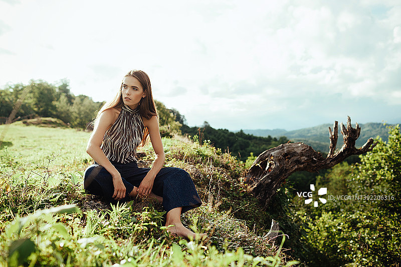 一位留着深色长发的年轻欧洲女子坐在悬崖边的草地上眺望远方图片素材