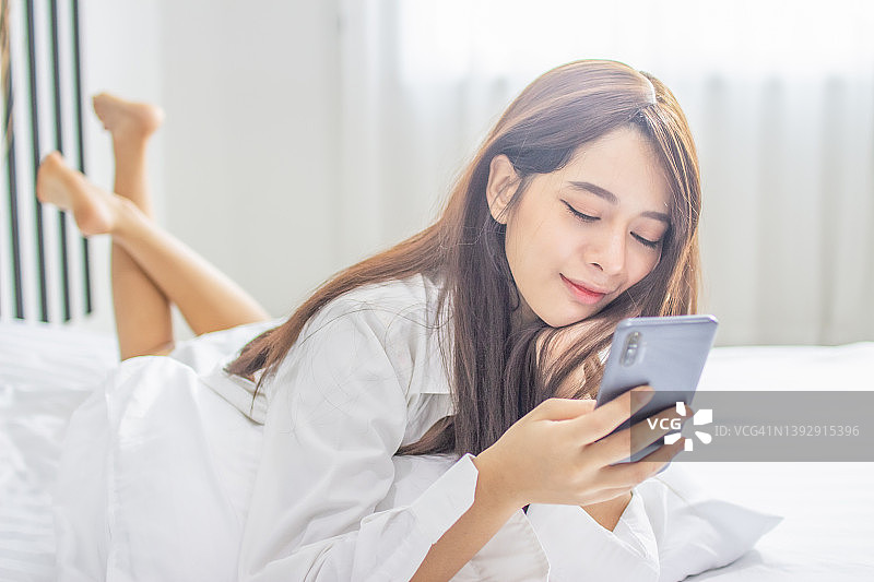 肖像年轻微笑快乐美丽的亚洲女人放松卧室。女人放松使用智能手机小玩意网上购物在家工作。图片素材