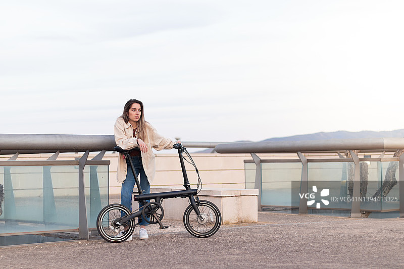 年轻女子靠在电动自行车看地平线等待她的朋友在城市公园在下午图片素材