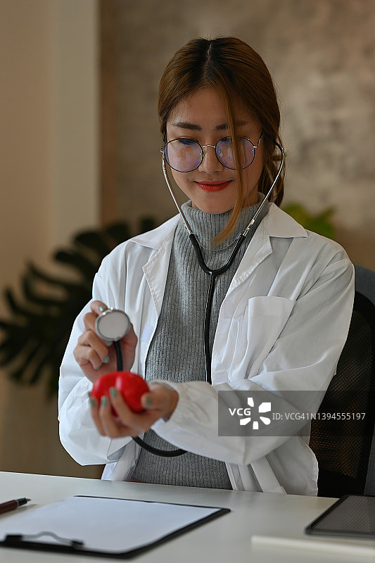 一位穿着白大褂的漂亮亚裔医生的肖像，坐在办公室里，手握心脏模型，面带自信的微笑，用听诊器做着检查姿势。为医疗保健理念。图片素材