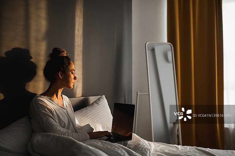 年轻的自由职业女性早上在床上用笔记本电脑工作。在家远程工作。图片素材