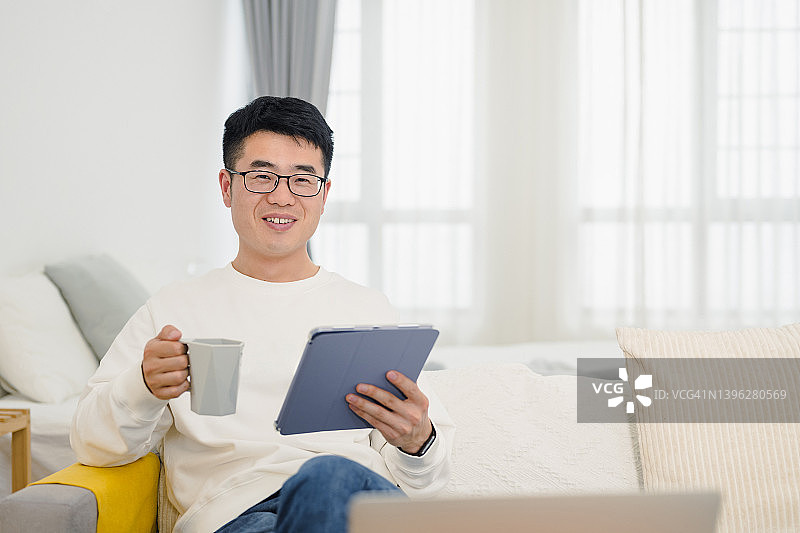 一名年轻人坐在客厅的沙发上，拿着咖啡杯使用平板电脑，看着镜头图片素材