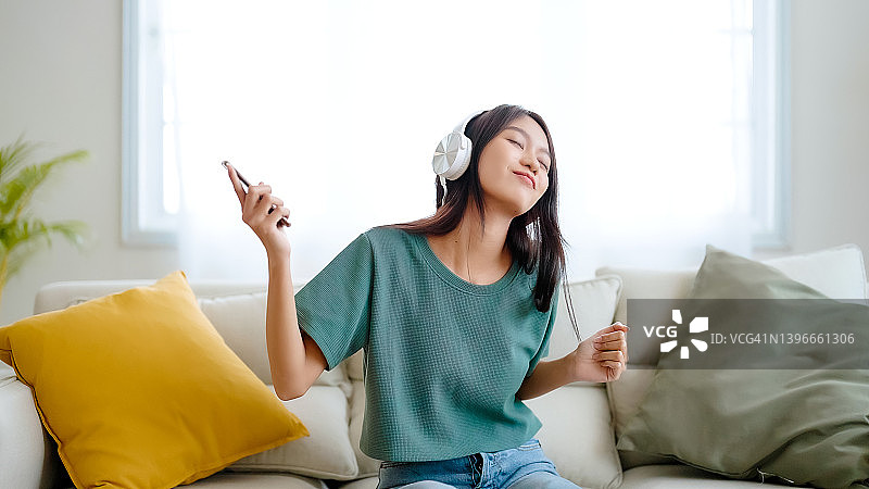 年轻的亚洲女性在家里客厅的沙发上听音乐。快乐亚洲女性使用手机、戴耳机、坐在沙发上图片素材