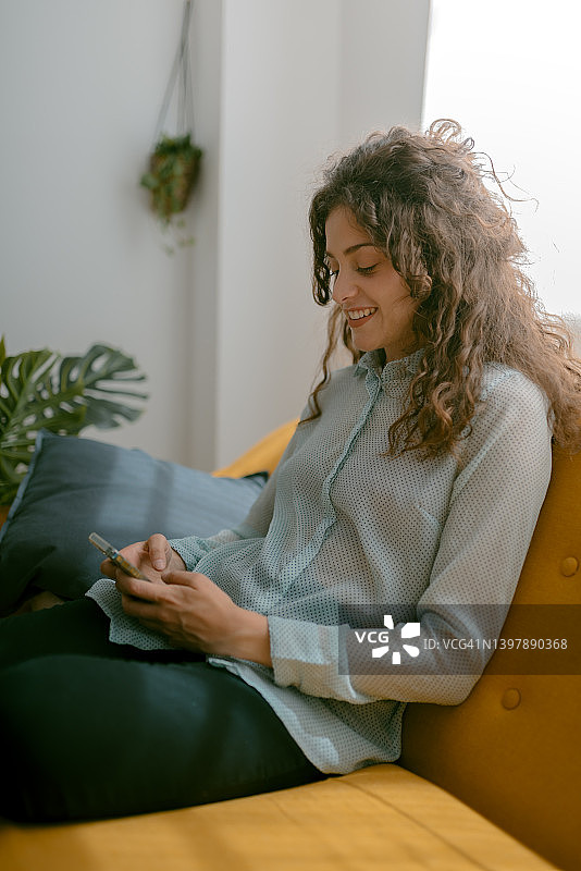 一头卷发的年轻女子坐在家里的沙发上，一边玩手机一边放松。图片素材
