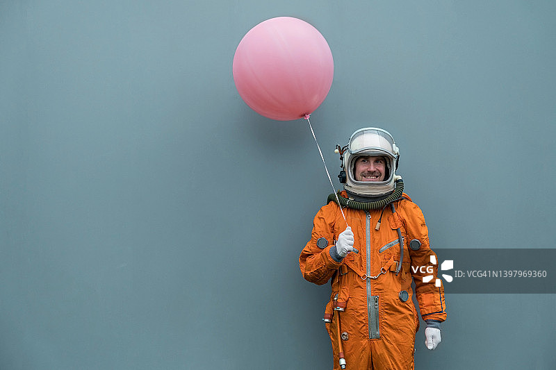 宇航员穿着太空服和头盔，拿着粉红色的充气气球，对着灰色的墙壁背景。快乐的宇航员和氦气气球图片素材