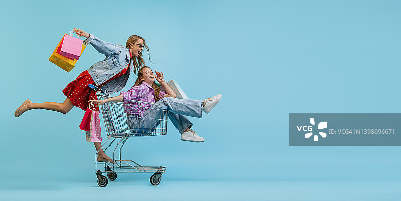 传单与漂亮快乐的女孩与购物袋骑在一个购物车孤立的蓝色背景。销售概念，黑色星期五，折扣，情感，青春和时尚图片素材