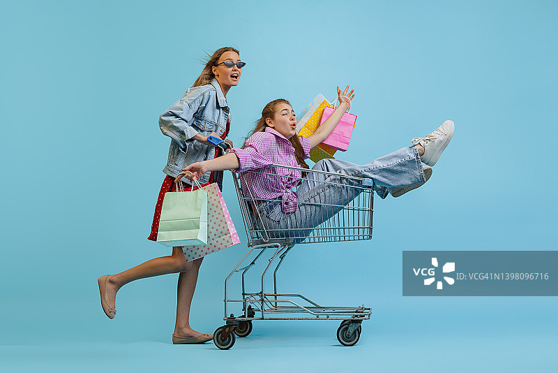 工作室拍摄的漂亮快乐的女孩与购物袋骑在一辆车孤立的蓝色背景。销售概念，黑色星期五，折扣，情感，青春和时尚图片素材