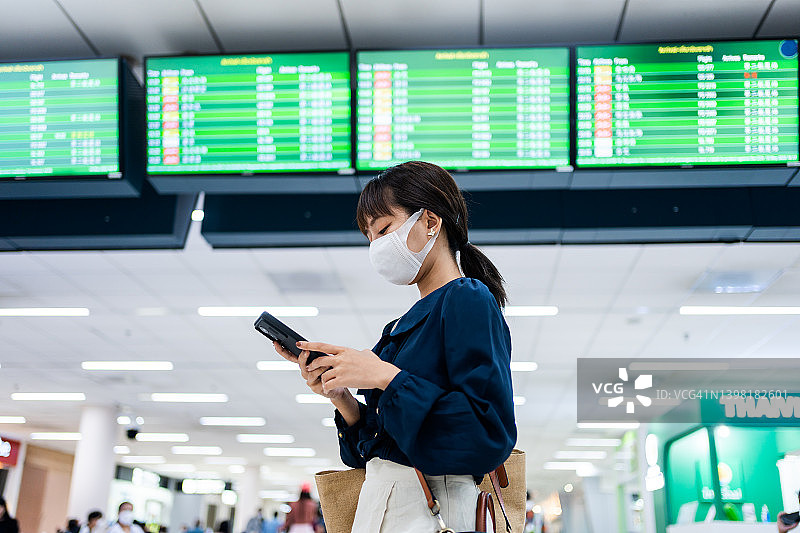在机场候机楼，一名亚洲年轻女子在飞机时刻表前使用智能手机图片素材