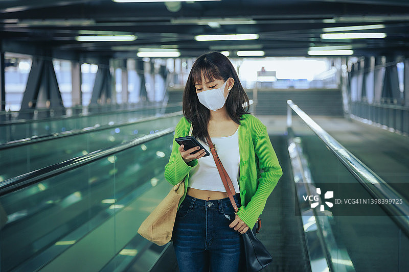 年轻的亚洲女商人戴着口罩，在机场的自动扶梯上用手机阅读电子邮件。商务旅行的概念。图片素材