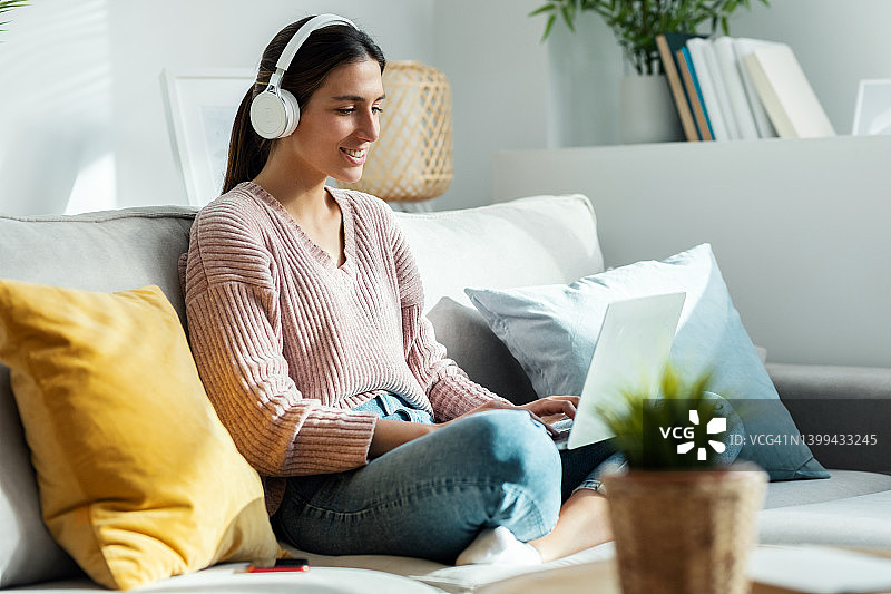 积极的年轻女性坐在家里的沙发上，一边用笔记本电脑听音乐，一边唱歌跳舞。图片素材
