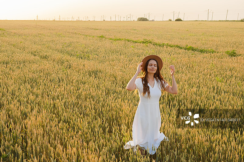 年轻美丽的千禧一代女子身穿白色长裙，帽子一顶，站在夕阳下金色的麦田里，眺望远方。图片素材