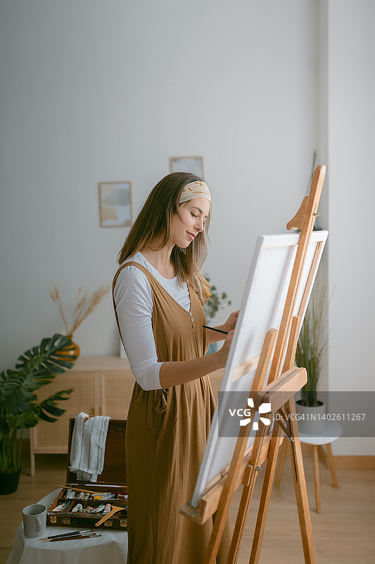才华横溢的女艺术家站在画室的木制画架前，在画布上作画。图片素材