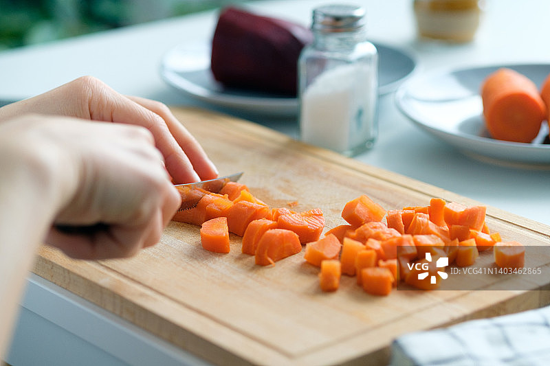 一个女人把煮熟的胡萝卜切成丁。准备一种叫做油醋汁的蔬菜沙拉。菜刀放在菜板上。附近还有其他食材。素食和纯素食品的概念。图片素材