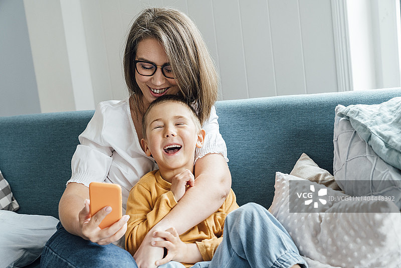 快乐男孩和妈妈抱着智能手机坐在家里的沙发上图片素材