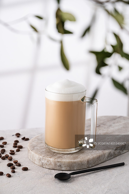 卡布奇诺或拿铁与牛奶泡沫在一个杯子与咖啡豆和勺子在光的背景与树枝和早晨的阴影图片素材