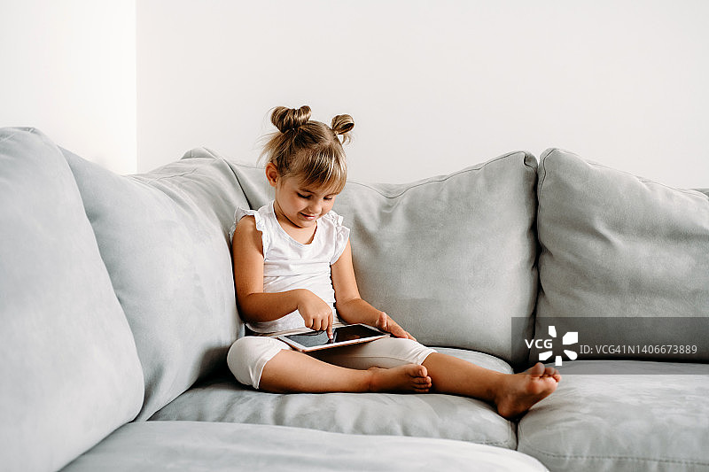 学步的小女孩在家里的沙发上玩数字无线平板电脑。宝宝的孩子成长与在线应用程序。儿童和电子设备的概念。婴儿肖像与智能手机。教育学习理念。图片素材