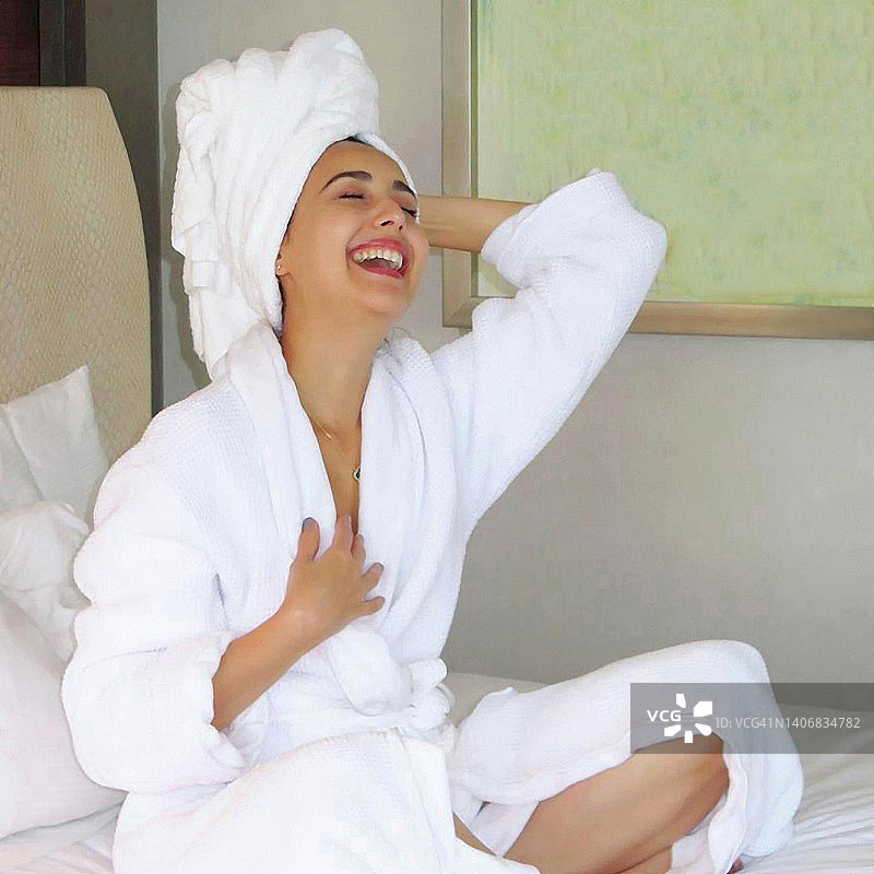 女人笑着坐在床上洗澡后，穿着浴袍和毛巾。图片素材
