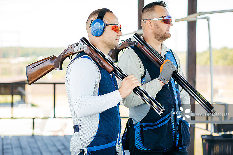 两名成年男子的肖像，戴着太阳镜，防护耳机和步枪背心练习射击武器。图片素材