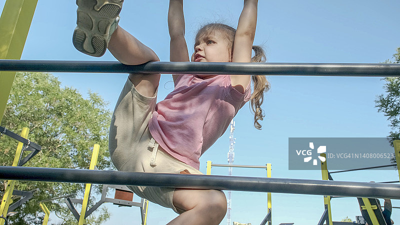 小女孩在户外运动场上爬体操梯。阳光灿烂的日子里，可爱的小女孩爬上城市公园里的垂直运动梯。图片素材