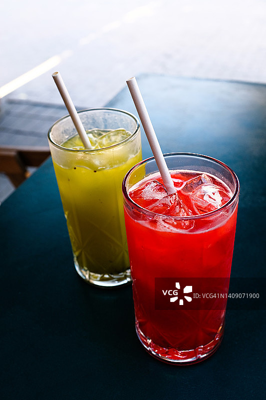 草莓浆果软饮料或加冰鸡尾酒，用吸管喝在玻璃杯子在户外咖啡馆或餐厅的木桌上。在炎热、闷热的夏天喝的清凉饮料。图片素材