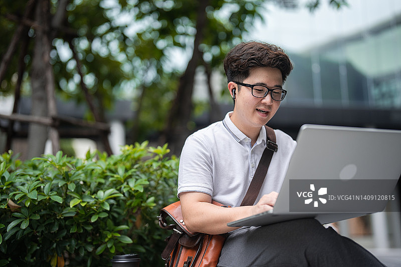 年轻的亚洲商人坐在城市公园的长凳上，喝着咖啡，在一天的工作中使用笔记本电脑工作。图片素材