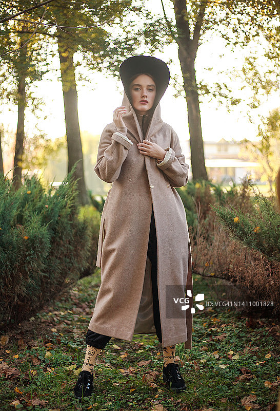 时尚时尚的年轻女子穿着米色大衣和连帽斗篷，背景是公园里的树，美女肖像的户外图片素材