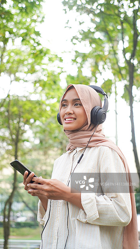 快乐的亚洲穆斯林妇女戴着头巾，在户外用智能手机和耳机听音乐图片素材