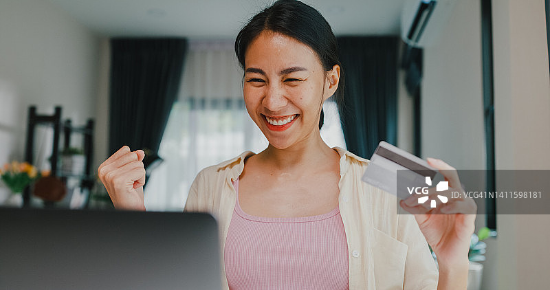 特写青年亚洲女女孩穿着休闲坐在书桌前与笔记本电脑持有信用卡感觉高兴和兴奋成功支付网上购物在客厅在房子。图片素材