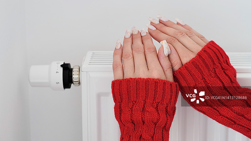 在白色墙壁的背景下，一个穿着红色毛衣的女人在电池上取暖的特写。微距照片的女人的手，复制空间。冬季节气理念。欧洲天然气危机。图片素材