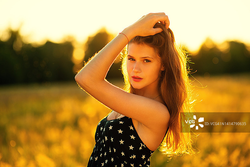可爱的年轻女子，长发穿着，微笑着站在草地上，在大自然的夕阳背景图片素材