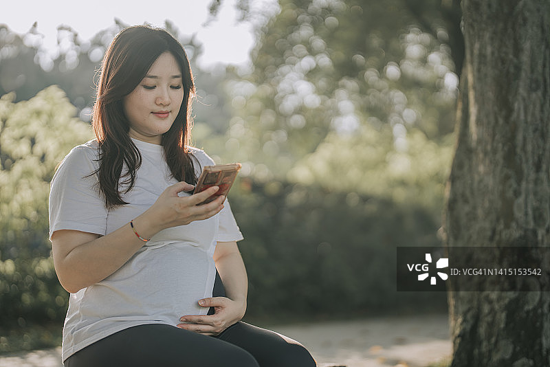 亚洲华人孕妇在公园坐在长椅上看智能手机图片素材