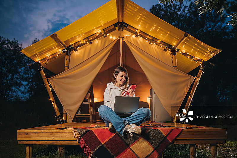 快乐女人自由职业者，夏夜在舒适的露营帐篷里用手机和笔记本电脑工作。用于户外度假和度假的豪华露营帐篷。生活方式的概念图片素材