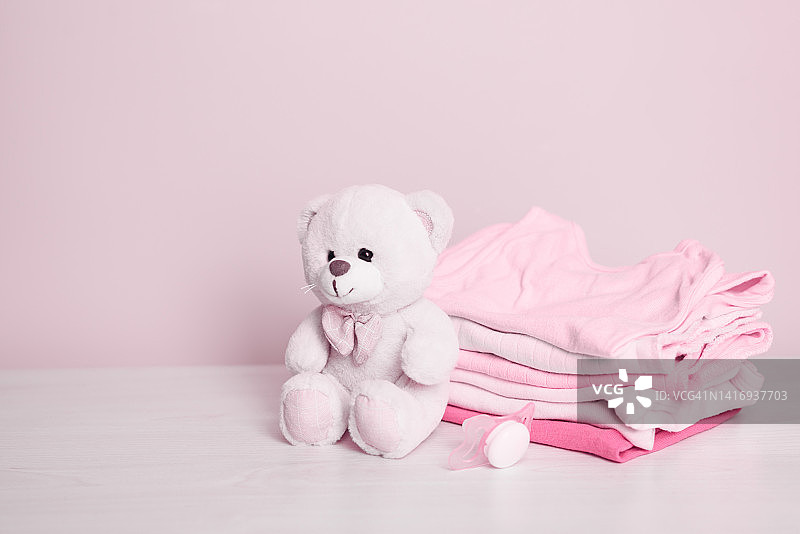 粉红色的背景上是一只小泰迪熊和一堆新生儿的干净衣服。复制文本空间。幼稚的概念。有选择性的重点。图片素材
