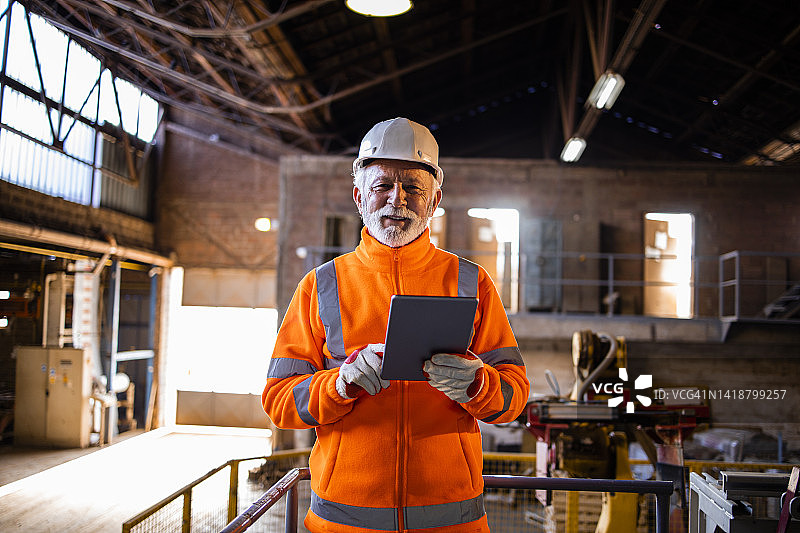 身穿橙色高能见度背心、头戴安全帽的工厂工人肖像，通过平板电脑控制生产过程。背景工业机械和内部。图片素材