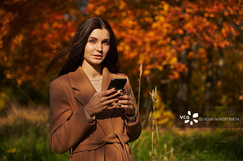 美丽的年轻女子在美丽的秋树的背景下，在阳光下用智能手机图片素材