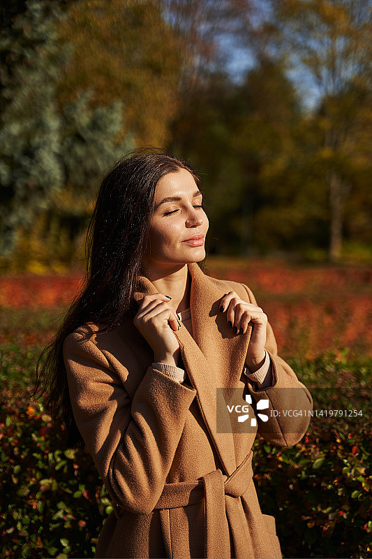 秋天的公园里，一位穿着大衣的美丽的年轻女子把手放在衣领上，面朝阳光，享受着阳光的光芒图片素材