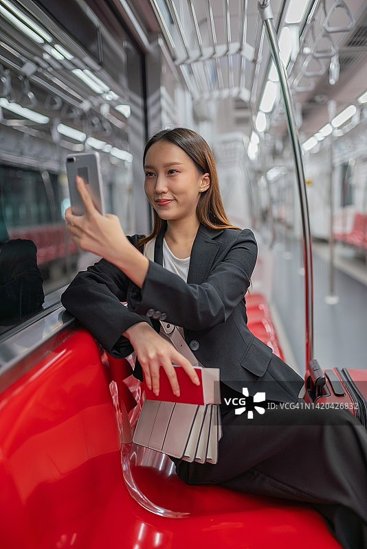 一名年轻的亚洲女商人的肖像照片旅行在通勤火车和红色行李与她的朋友视频通话，并在旅途中为商务旅行在她的智能手机自拍图片素材
