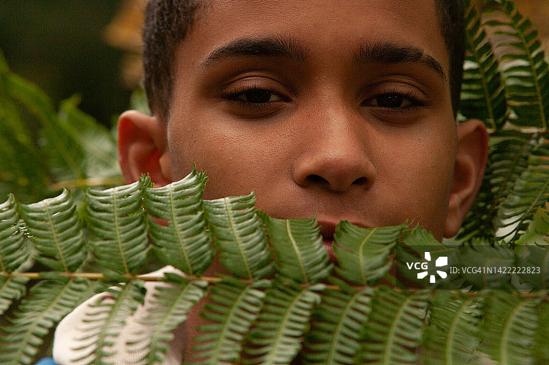 风景方向的一个英俊的年轻人与深色船员剪藏在一个甘美的绿色蕨类叶子后面，一个十几岁的男孩在大自然中看着相机与他的脖子和他的嘴都被树叶隐藏图片素材