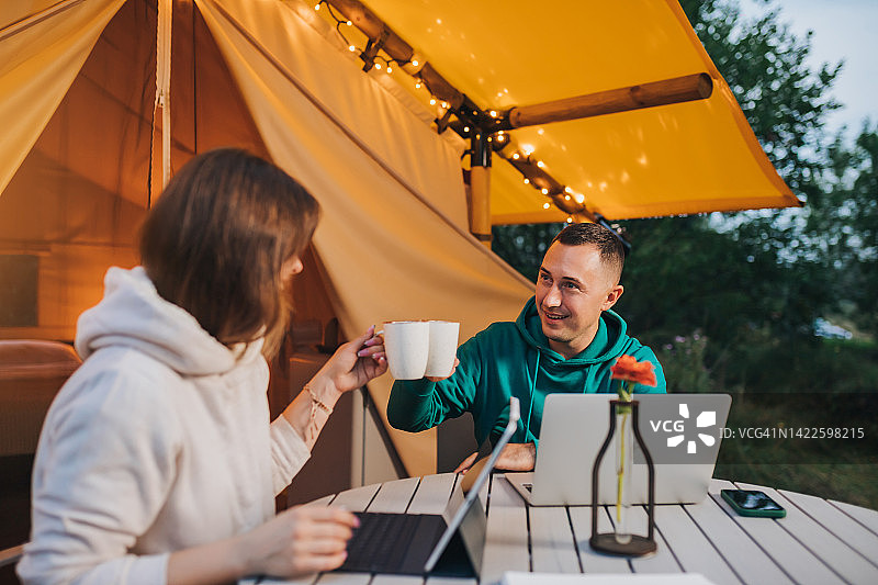 快乐的家庭夫妇自由职业者喝着咖啡，在舒适的野营帐篷里用笔记本电脑。户外度假度假的豪华露营帐篷。生活方式的概念图片素材