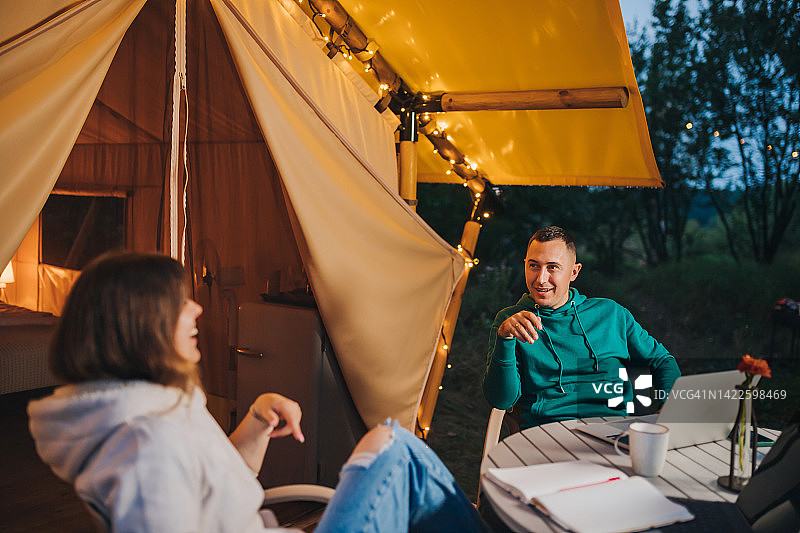 快乐的家庭夫妇自由职业者在舒适的野营帐篷里用笔记本电脑休息。户外度假度假的豪华露营帐篷。生活方式的概念图片素材