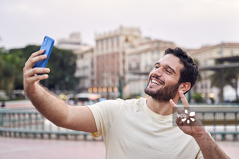 正面的一个微笑的年轻人在城市用他的手机自拍。图片素材