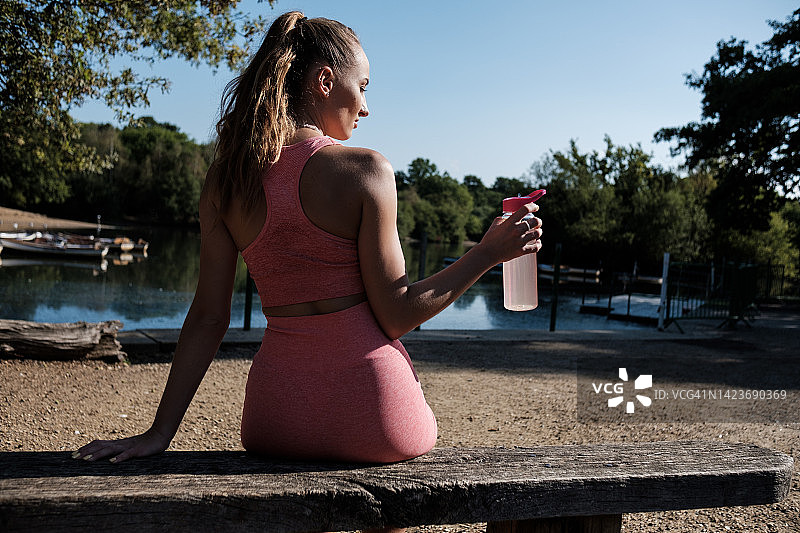 一位年轻的妇女正在锻炼后坐在长凳上休息，正在喝水。图片素材