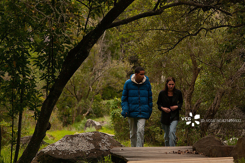 一对年轻人在绿色地带散步和聊天的景观朝向，两个穿着随意的牛仔裤的青少年沿着木制板条步道走向相机，同时沉浸在大自然中绿叶绿色地带的谈话中图片素材