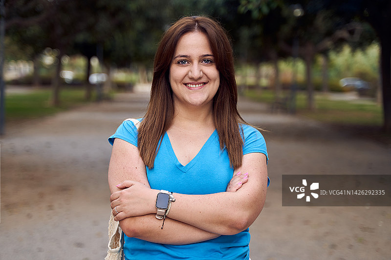 正面特写的肖像，一个快乐的年轻女子穿着蓝色t恤，双臂交叉在城市公园。图片素材