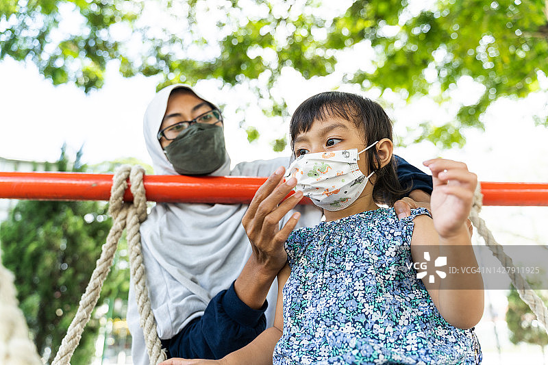 亚洲年轻母亲帮助女儿戴上防护口罩在操场上玩耍图片素材