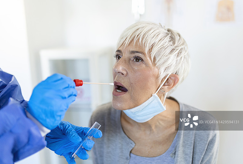 一位成熟的白人女性在临床环境中被穿着防护服的医护人员拭子，以确定她是否感染了冠状病毒或COVID-19。图片素材