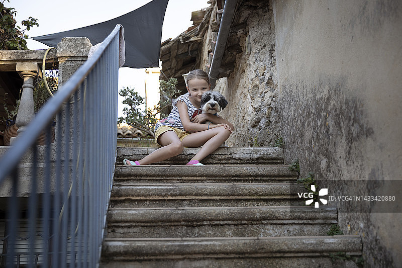 夏天，女孩(8-9岁)坐在度假别墅的石阶上，抱着一只宠物狗。图片素材