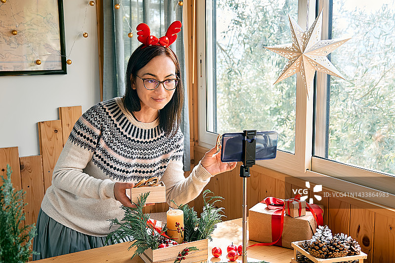 女花匠博主在她的智能手机前用新鲜的松枝、蜡烛和圣诞装饰制作冬季花道。小生意。季节性冬季车间。图片素材