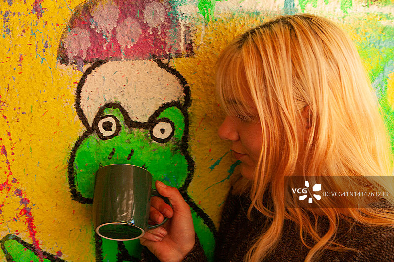 一个快乐的美丽的年轻女子拿着一个绿色的咖啡杯在一个彩色的墙壁上画青蛙，一个微笑的少女长着金色的头发和刘海拿着她的杯子在一个充满活力的绿色青蛙与红色蘑菇作为帽子在另一个黄色的背景图片素材
