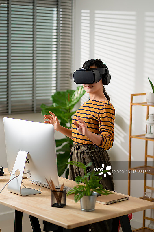 亚洲女性戴着VR头戴设备，在空中触摸，在家里办公室微笑。图片素材